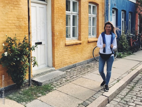 Pretty Caucasian lady with long hair, wearing jeans, white shirt and blue jacket on shoulders, smiling at camera, talking a walk along colorful buildings. Traveling and street style concept.