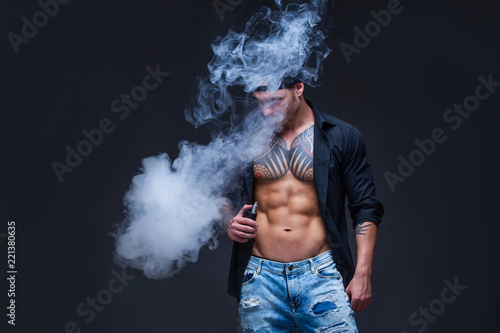 Vaper. The man dressed blue jeans, black shirt and black baseball cap with tattoos smoke an electronic cigarette on the dark background