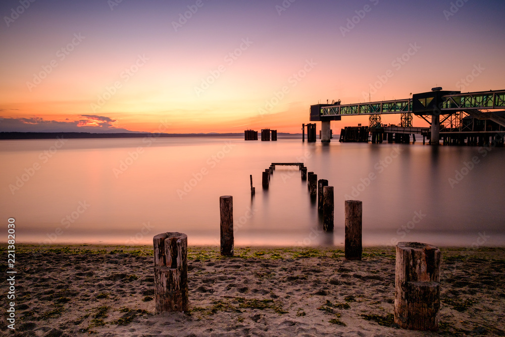 Long exposure of pilings leading in to a calm sea at sunset, with a ferry terminal in background