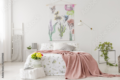A bunch of yellow fresh cut flowers in a bright bedroom interior with a bed dressed in white linen and peach blanket. Fabric on the wall above the bed. Real photo. photo