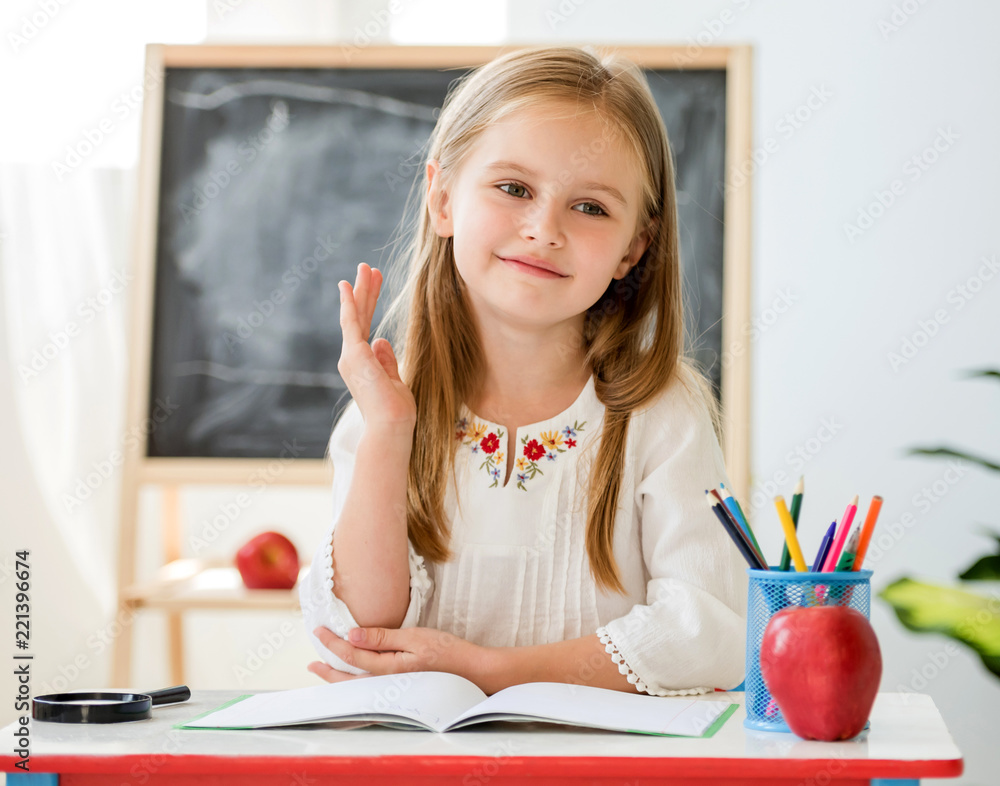 Little blond girl raise a hand for the answer sitting at the desk in ...