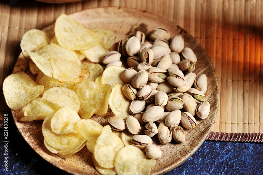 Pistachios and potato chips on a wooden plate. Under the plate is a light bamboo napkin. Close-up. View from above. Pistachios and potato chips on a wooden plate. Under the plate is a light bamboo napkin. Close-up. View from above.