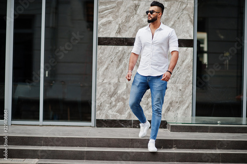 Stylish tall arabian man model in white shirt, jeans and sunglasses posed at street of city. Beard attractive arab guy.