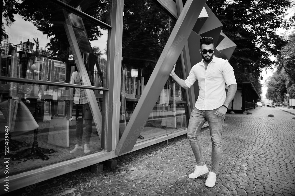 Stylish tall arabian man model in white shirt, jeans and sunglasses posed at street of city. Beard attractive arab guy.