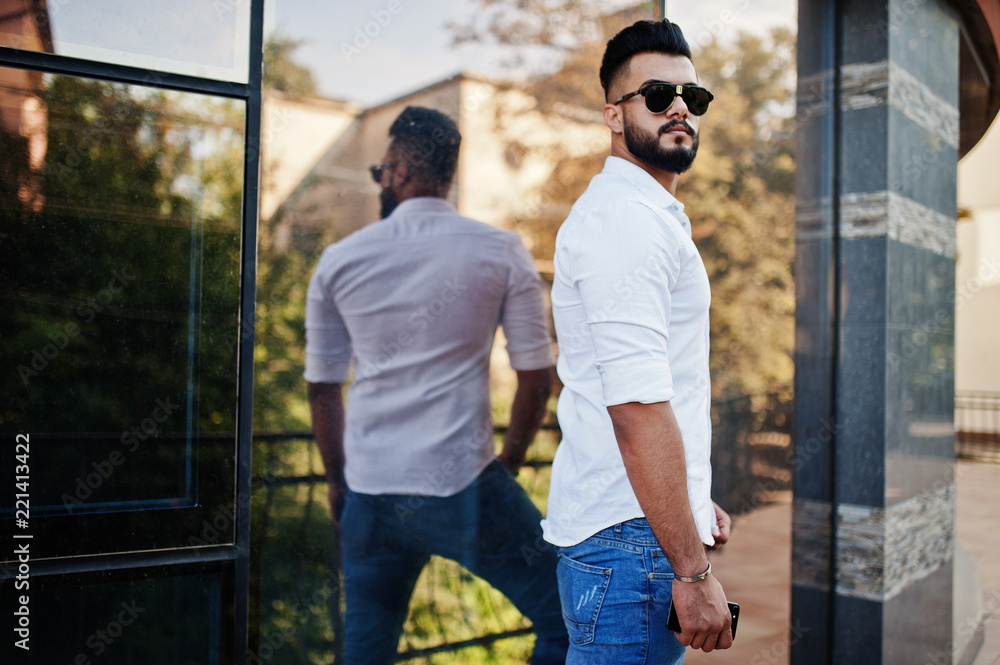 Stylish tall arabian man model in white shirt, jeans and sunglasses posed at street of city. Beard attractive arab guy against modern building. Stylish tall arabian man model in white shirt, jeans and sunglasses posed at street of city. Beard attractive arab guy against modern building.