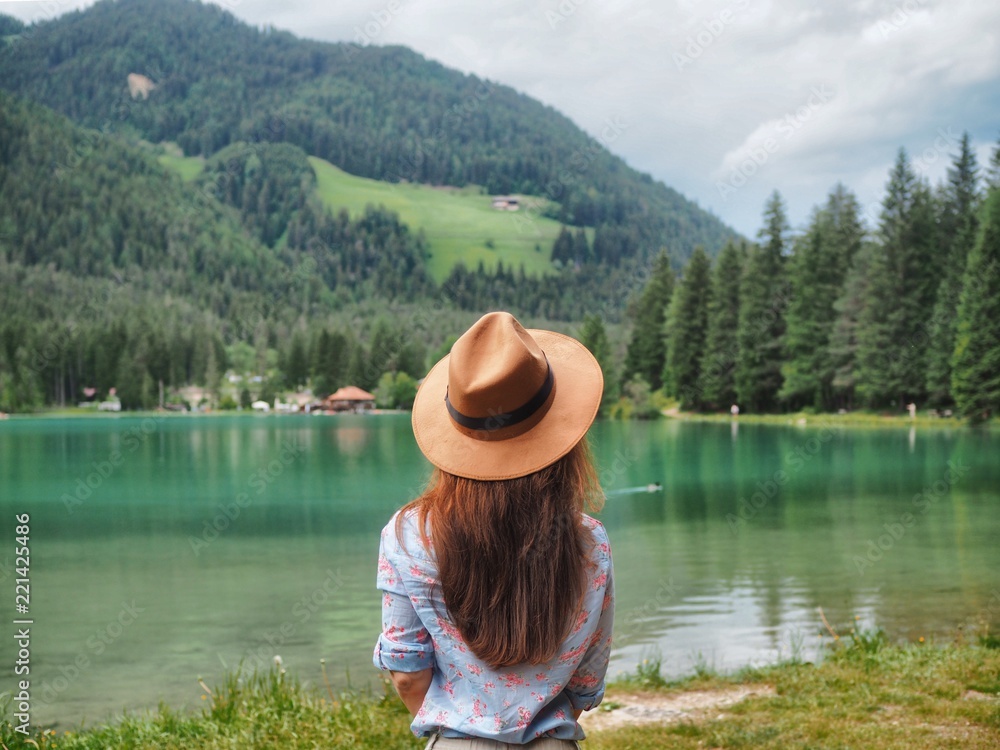 A girl with long hair and a shirt looking straight ahead at the azure mountain lake in the Dolomites, where the dense coniferous forests. The girl is wearing a brown wide-brimmed hat