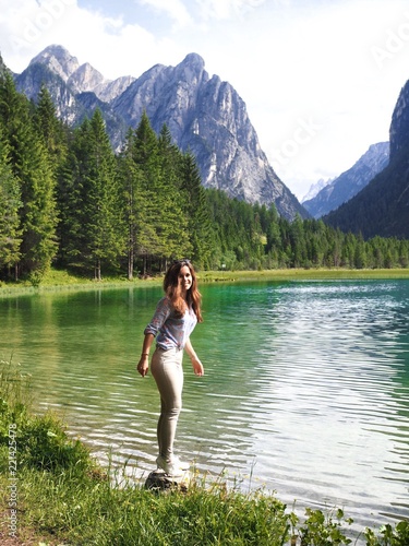 A girl with long hair and a shirt standing on a pebble near mountain azure lake in the Dolomites, where dense coniferous forests