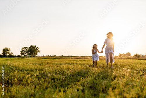 mother and daughter walking in green meadow with sunset on background