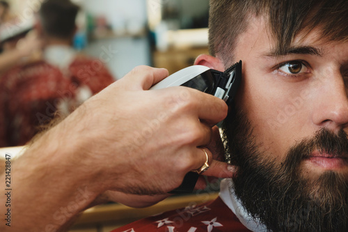 Handsome bearded man in barbershop. Barber cuts hair with  electric trimmer. photo