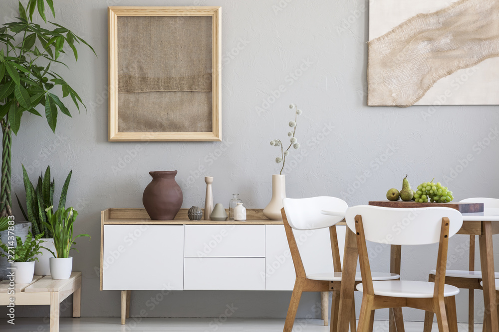 Dining table with fresh fruits and white wooden chairs in real photo of light grey room interior with two posters, green plants and small cupboard with decor Dining table with fresh fruits and white wooden chairs in real photo of light grey room interior with two posters, green plants and small cupboard with decor