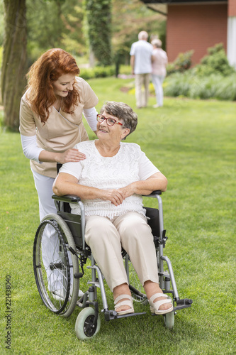 Billede på lærred A professional caretaker assisting and standing behind a senior woman patient on a wheelchair on a summer day in the garden of a private rehabilitation clinic
