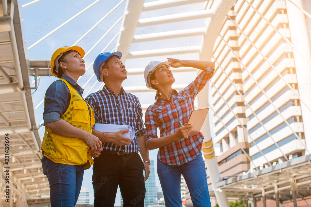 Obraz premium Three industrial engineer wear safety helmet and holding tablet engineering working and talking with drawings inspection on building outside. Engineering tools and construction concept.