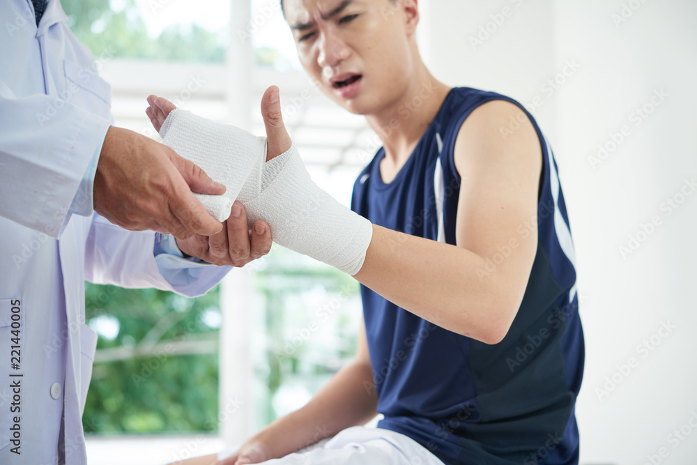 Anonymous medical practitioner applying clean bandage on injured arm of young Asian guy while working in office Anonymous medical practitioner applying clean bandage on injured arm of young Asian guy while working in office