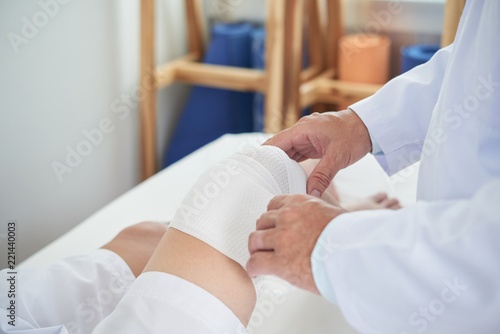Anonymous medical practitioner using clean bandage to dress knee of unrecognizable man on blurred background of doctor's office