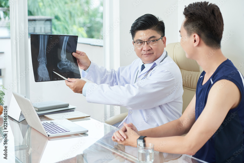 Φωτογραφία Adult Asian man in medical apparel demonstrating X-ray picture of legs to young