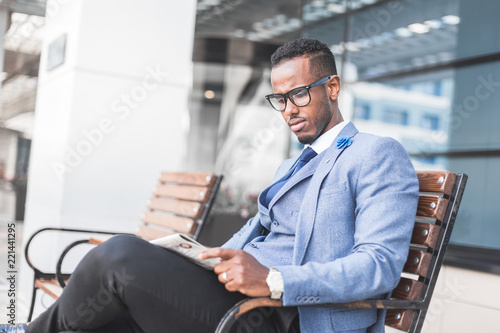 black man businessman in a business suit and glasses sits on a bench and reads a newspaper against the backdrop of a modern city