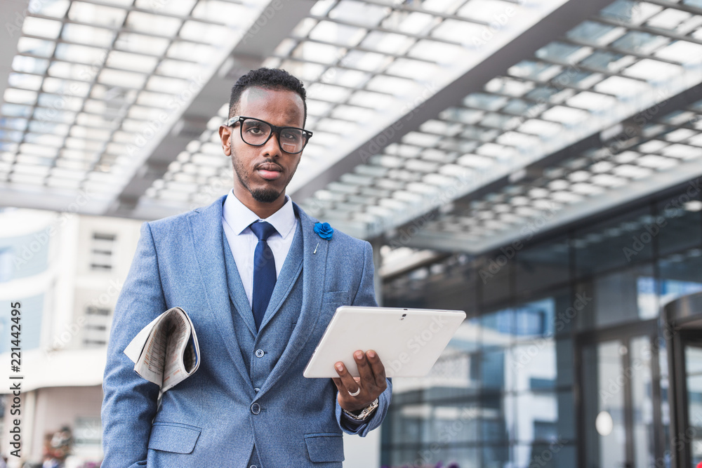 black man businessman in a business suit, expensive watch and glasses with a newspaper and digital tablet goes against the backdrop of a modern city to work black man businessman in a business suit, expensive watch and glasses with a newspaper and digital tablet goes against the backdrop of a modern city to work