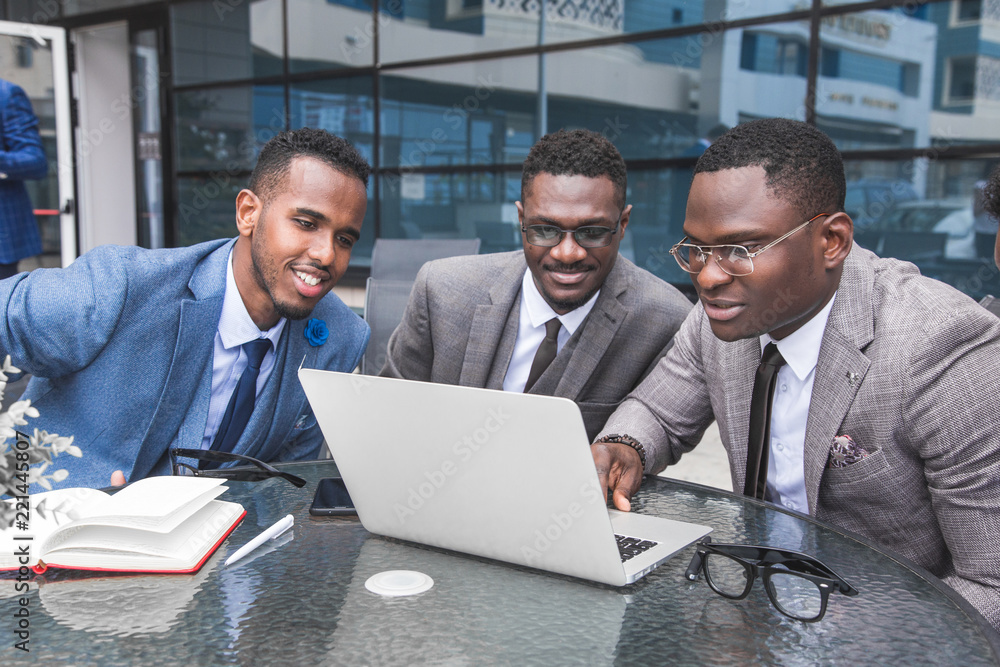 Group of happy diverse male and female business people team in formal gathered around laptop computer in bright office against the background of a glass building Group of happy diverse male and female business people team in formal gathered around laptop computer in bright office against the background of a glass building