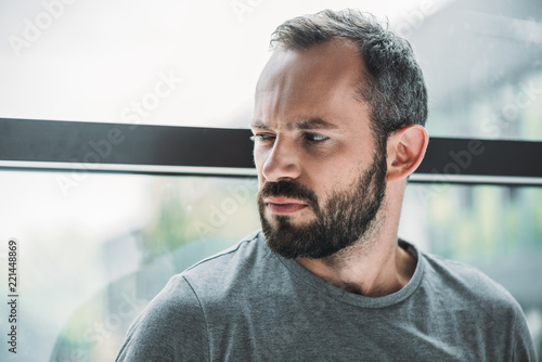 portrait of unhappy bearded man standing near window and looking away