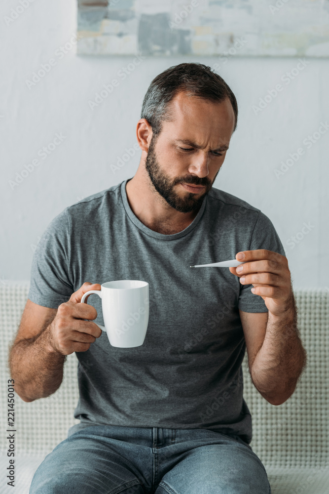 sick bearded middle aged man holding cup and looking at thermometer sick bearded middle aged man holding cup and looking at thermometer