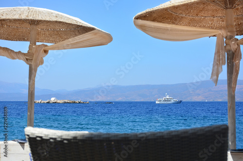 Fototapeta Naklejka Na Ścianę i Meble -  Small island with wedding chapel and ship in the sea visible through beach umbrellas from the shore in Hydra island, Greece