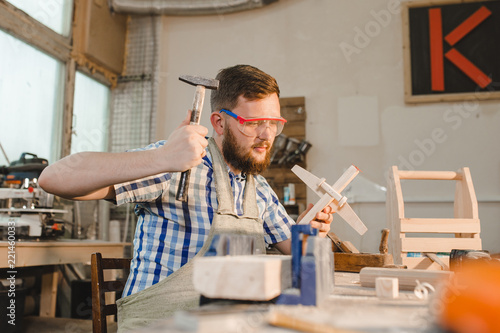 a bearded man in safety glasses and an apron makes a wooden plane in a carpentry workshop. The father makes a toy for his son with his own hands. Carpenter hammers a nail into a wooden plane. Toy work