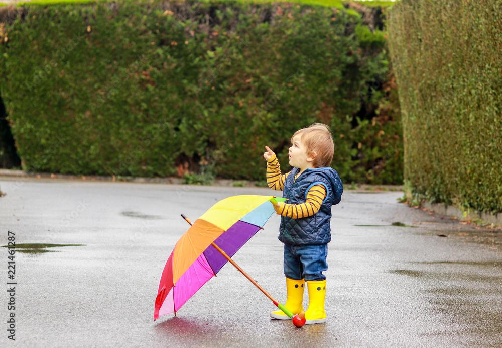 Cute little baby boy in yellow rubber boots with colorful rainbow umbrella  staying on wet road after rain pointing with his finger up at sky. Copy  space. Autumn kid fashion Stock Photo