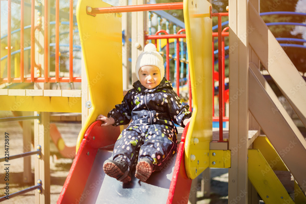 Smiling little boy in a black jumpsuit and white cap sitting on a children slide in a sunny day. Active childhood concept