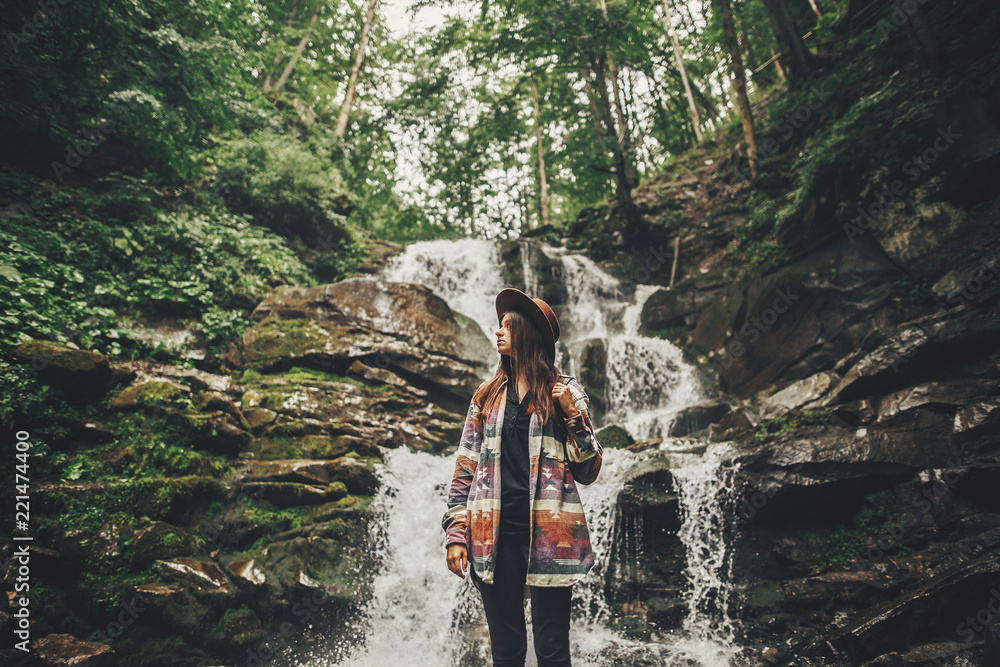 stylish hipster girl in hat with backpack standing at waterfall in forest in mountains. traveler woman exploring woods. travel and wanderlust concept. atmospheric moment stylish hipster girl in hat with backpack standing at waterfall in forest in mountains. traveler woman exploring woods. travel and wanderlust concept. atmospheric moment