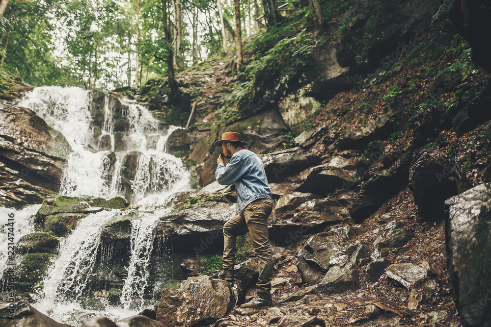 stylish hipster man in hat with photo camera, taking images of waterfall in forest in mountains. traveler guy exploring woods. space for text.  travel and wanderlust concept