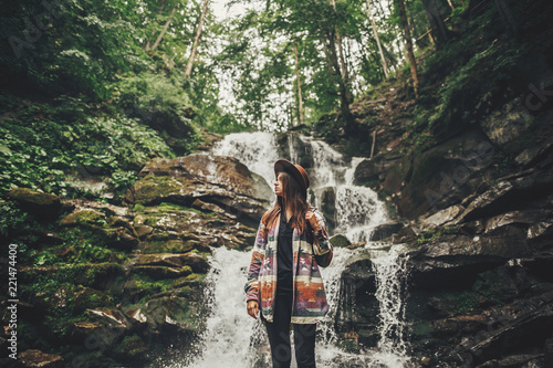 stylish hipster girl in hat with backpack standing at waterfall in forest in mountains. traveler woman exploring woods. travel and wanderlust concept. atmospheric moment