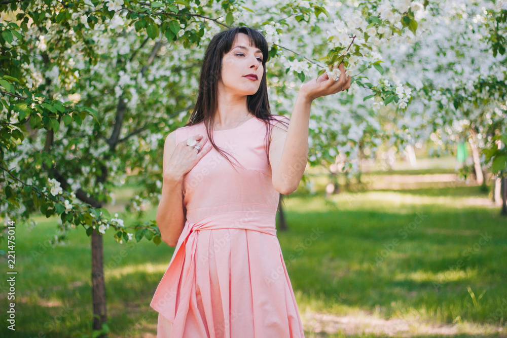 A beautiful girl with long black hair in a pink dress is walking in an apple orchard surrounded by flowers.. A model with clean skin. White flowers on the trees. A beautiful girl with long black hair in a pink dress is walking in an apple orchard surrounded by flowers.. A model with clean skin. White flowers on the trees.