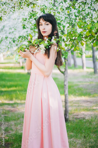 A beautiful girl with long black hair in a pink dress is walking in an apple orchard surrounded by flowers.. A model with clean skin. White flowers on the trees.