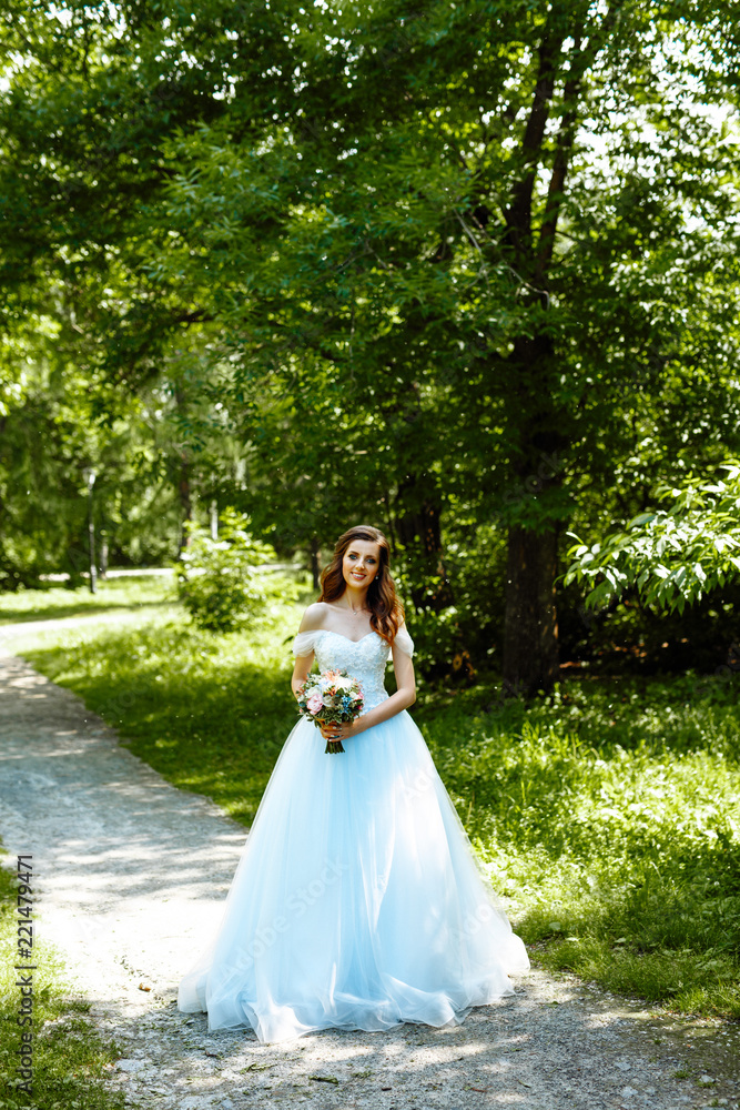 Portrait of beautiful brunette bride with curly hair in Park on green foliage background. Girl in a lush blue wedding dress with a bouquet of flowers.