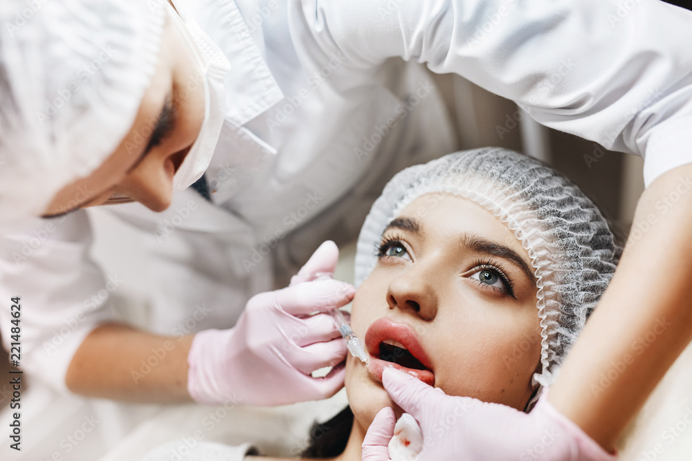 The process of lips enhancement. Cosmetician makes an injection of hyaluronic acid in a beautiful lips. The young girl with a beautiful face in the special hat and hands of the doctor in the pink glov The process of lips enhancement. Cosmetician makes an injection of hyaluronic acid in a beautiful lips. The young girl with a beautiful face in the special hat and hands of the doctor in the pink glov
