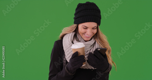 Smiling woman in winter clothes with coffee texting on phone on green screen