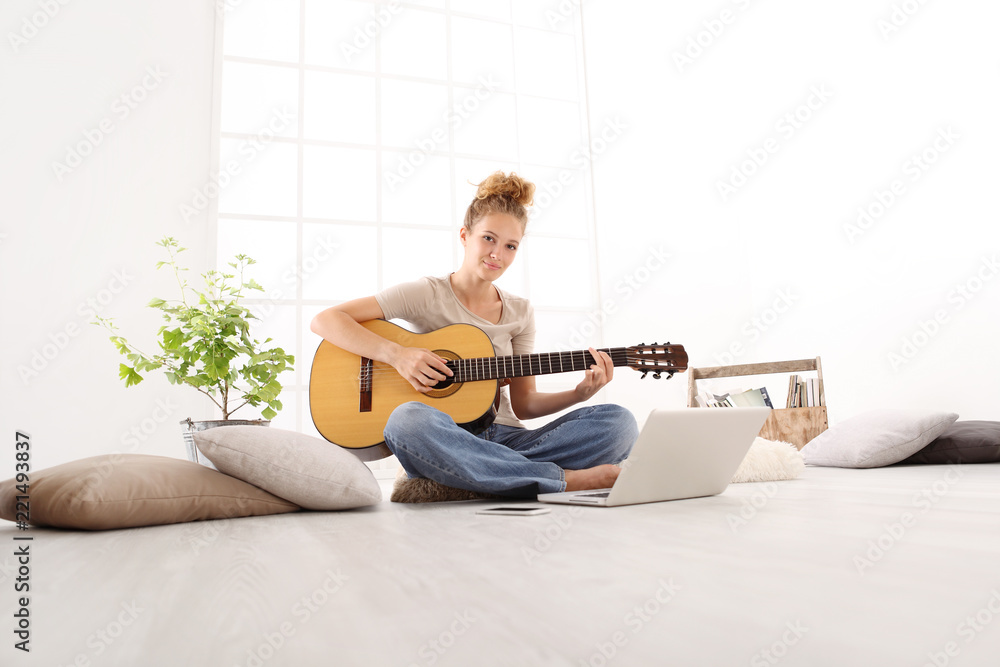 Obraz premium Beautiful smiling young woman playing guitar with computer, learn to play with an online course, sitting on the floor in casual clothes at home