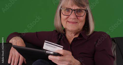 Charming old woman with tablet and credit card smiling at camera on green screen