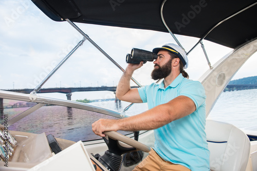 A young guy with a beard sails on a yacht at the helm with binoculars in his hands