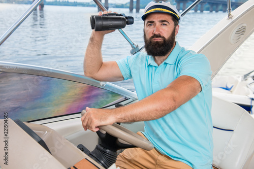 A young guy with a beard sails on a yacht at the helm with binoculars in his hands