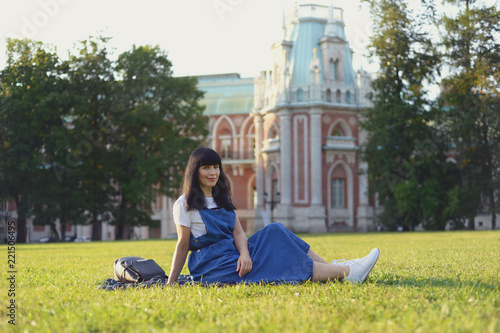 Stylish woman in a jeans dress resting on the green grass in the park. She sits in a clearing, behind a beautiful palace. Warm sunny day. Moscow. Tsaritsino. photo