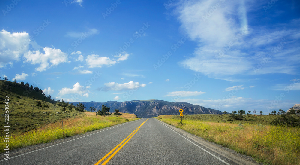Green hills dotted with small trees and mountains divided by asphalt highway with a speed limit sign under a sunny blue sky in a summertime Wyoming landscape