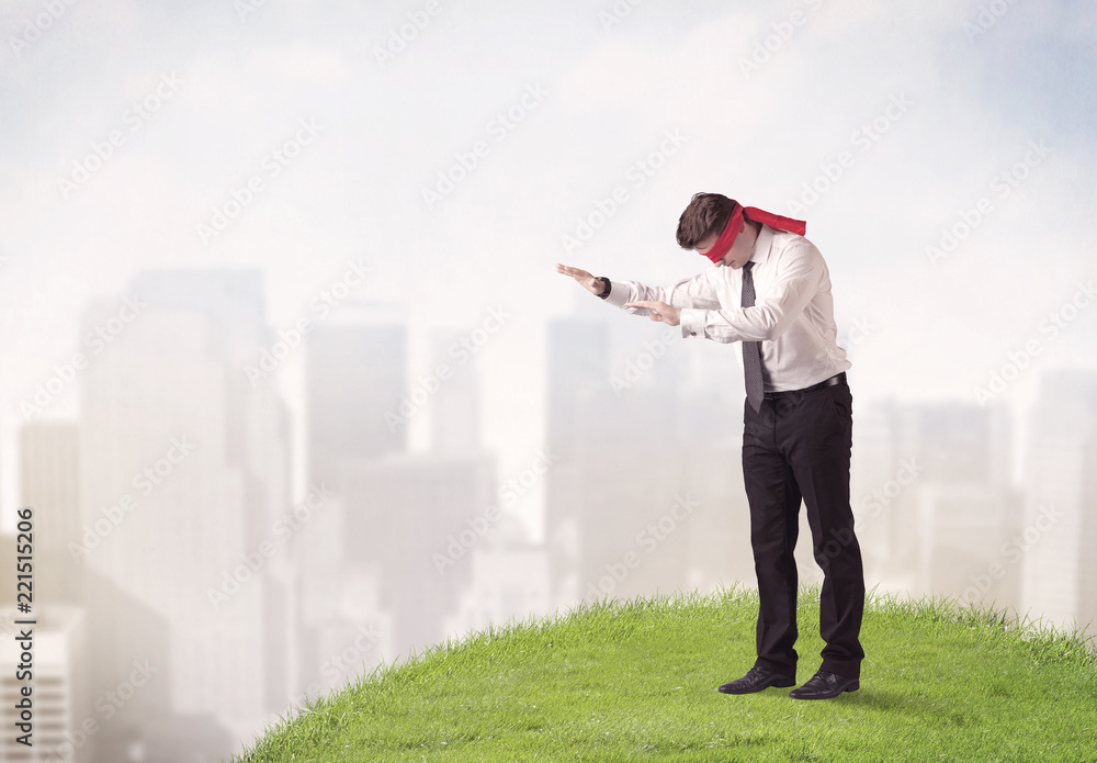 Young blindfolded businessman steps on a a patch of grass with a city in the background Young blindfolded businessman steps on a a patch of grass with a city in the background