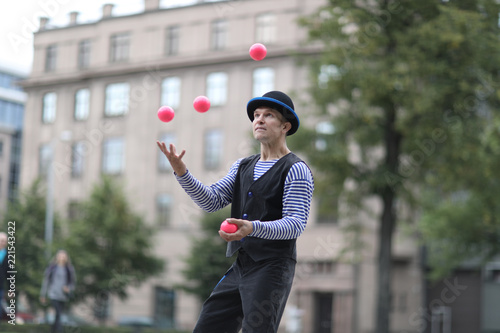 A male clown juggles with red balls in the town square