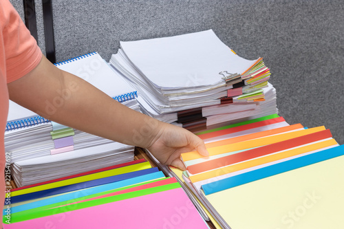 Student is sending homework assignment on teacher's table, which has a large number of paperwork stacked in archive with colorful paper clips and plastic binding bars. Education and business concept.
