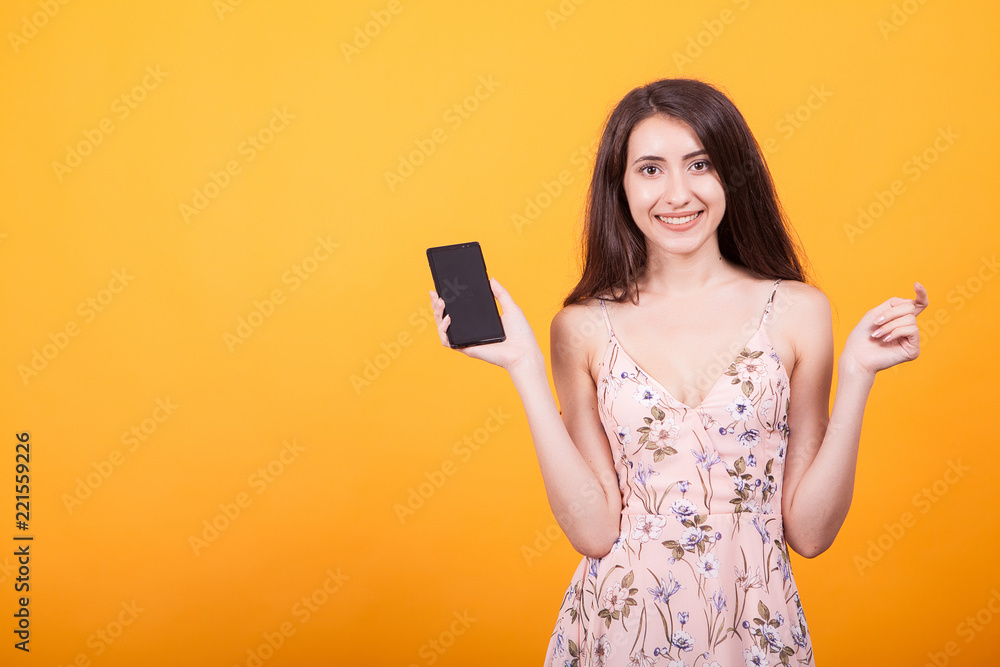 Naklejka premium Cute young woman pointing on her phone in studio on yellow background.
