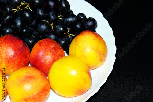 Peaches  black seedless grapes and nectarines. Orange juice and fruit for Breakfast. Yellow tropical fruit on a white plate. A glass of juice on a black background. Vegetarian and raw food menu. Diet