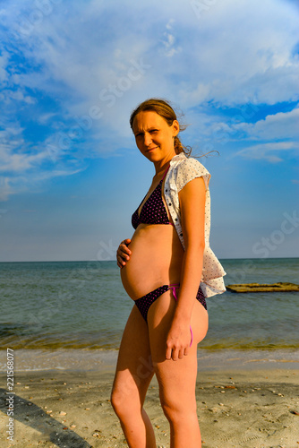 Fototapeta Naklejka Na Ścianę i Meble -  A pregnant woman in a white blouse is standing on the beach.