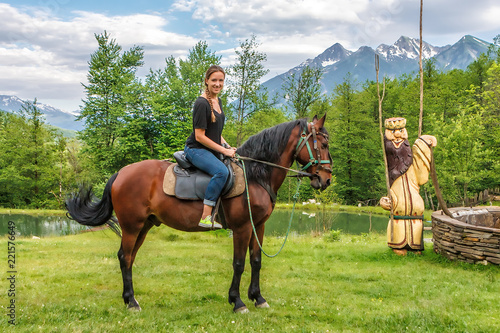 Young happy smiling Caucasian woman wearing jeans rides brown horse posing and smiling at camera in Caucasus mountain countryside on snowy Aibga peaks background in summer