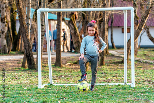 Young Caucasian goalkeeper girl with flushed face posing by outdoor football goal with foot put on ball looking into camera with frowning glance and slight smile